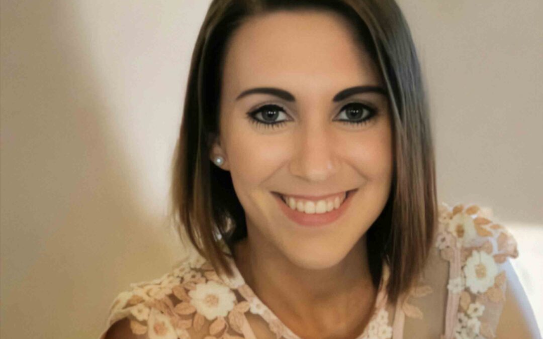 Lächelnde Frau mit schulterlangem braunem Haar und heller, floraler Spitzenbluse vor einem neutralen, weich ausgeleuchteten Hintergrund.