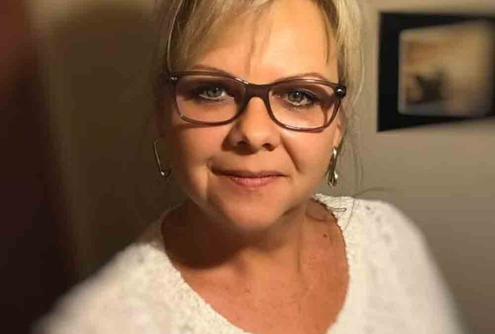 Porträt einer lächelnden Frau mit kurzer blonder Frisur und Brille. Sie trägt einen weißen Pullover und steht in einem Innenraum mit warmem Licht und unscharfem Hintergrund.