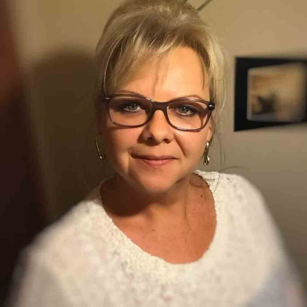 Porträt einer lächelnden Frau mit kurzer blonder Frisur und Brille. Sie trägt einen weißen Pullover und steht in einem Innenraum mit warmem Licht und unscharfem Hintergrund.